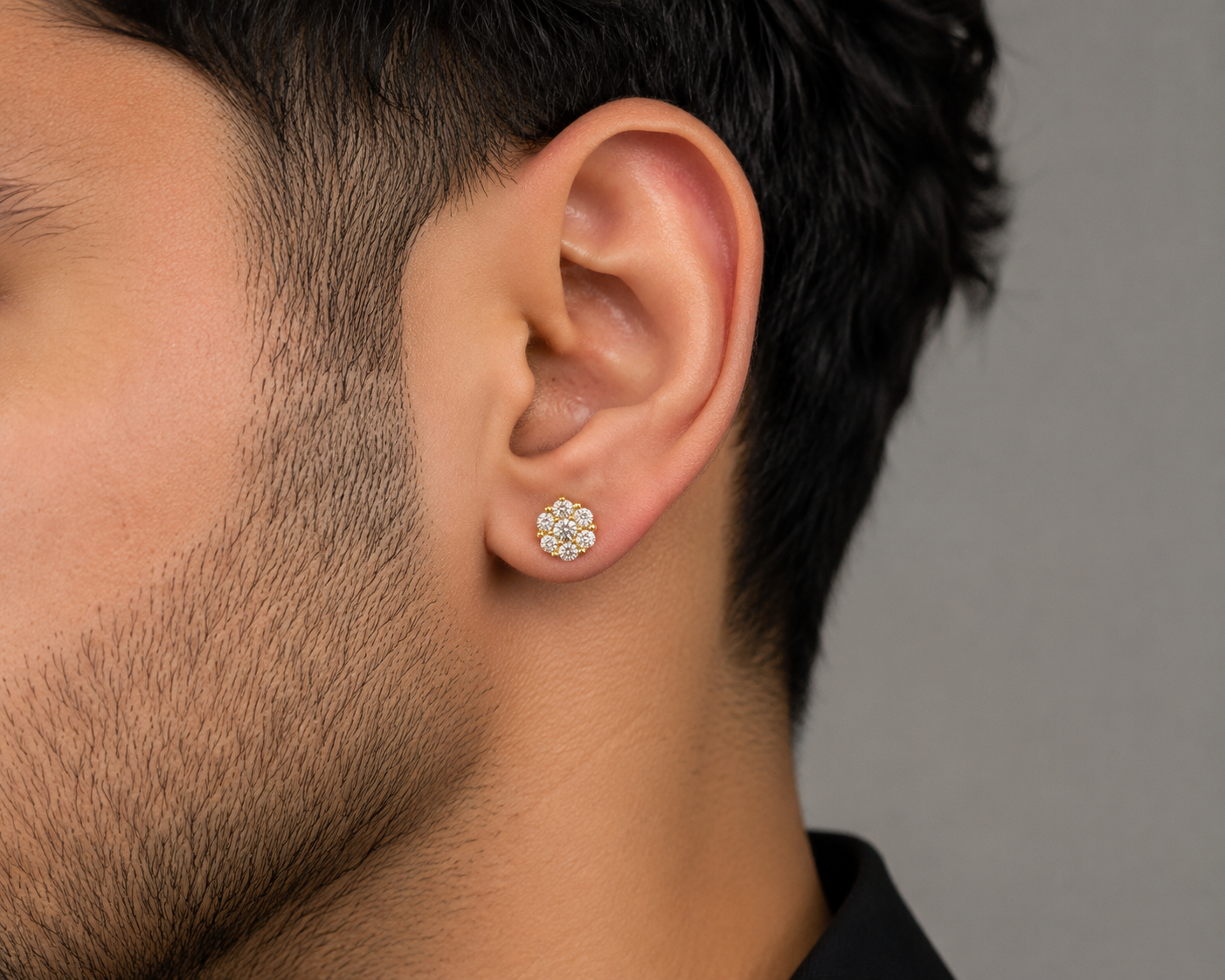 A close-up side profile of a man's ear wearing a small gold flower-shaped stud earring with clear gemstones, set against a neutral gray background.