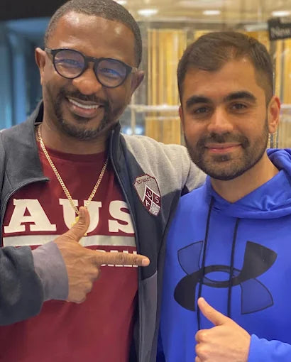 Two men posing together, one pointing at the other, with a blurred indoor background.